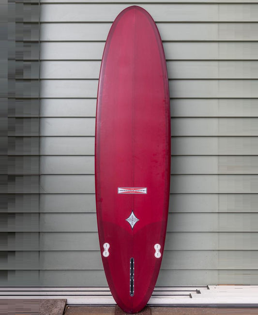 The Gordon & Smith G&S Mid length Cardinal 76 surfboard, featuring a rounded nose and fin boxes, stands upright against a gray horizontal panel wallโa versatile red board perfect for any quiver.