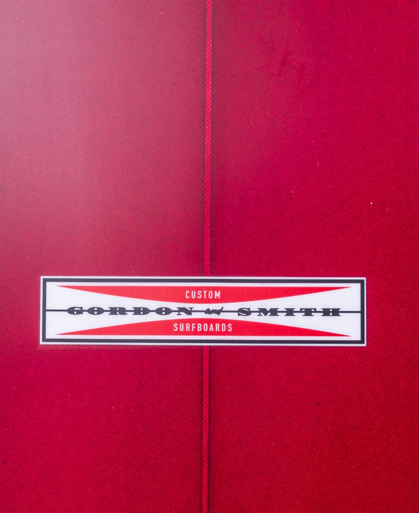 Close-up of a red G&S Mid length Cardinal 76 surfboard by Gordon & Smith, featuring a centered label with the brand’s name in black and red text on a white background.