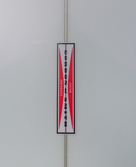 A rectangular red and white label reading Gordon & Smith evokes classic paddle power, displayed on a frosted glass surface alongside the G&S Long Twin Skylight 68 surfboard.