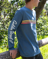 Side view of a person wearing a Heavy Island Long Sleeve Tee | Blue with retro stripe design across the chest and "Golden Breed" printed on the sleeve, outdoors in a sunny setting.
