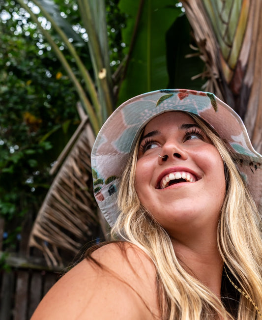 A woman with long blonde hair wears the Golden Breed Chelsea Bucket Hat in Rose and smiles while looking upward, with tropical plants in the background.