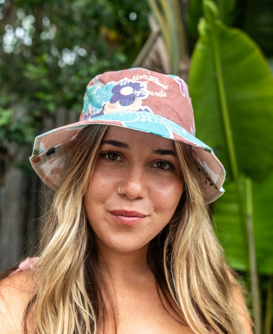 A woman with long blonde hair wears the Chelsea Bucket Hat | Rose by Golden Breed, standing outdoors amid lush green tropical plants.