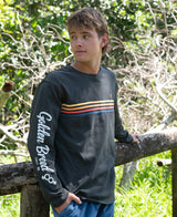 A young man wearing a Heavy Island Long Sleeve Tee | Wash Black with multicolored stripes across the chest and white script on the sleeve, standing outdoors near greenery and a wooden fence.