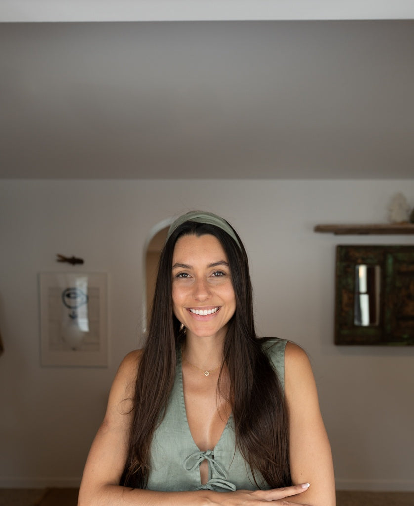 A woman with long brown hair, wearing a green sleeveless top and the Lazy Beach Head Scarf Linen in Dark Khaki, stands indoors smiling at the camera with her arms folded, framed art and a decorative shelf in the background.