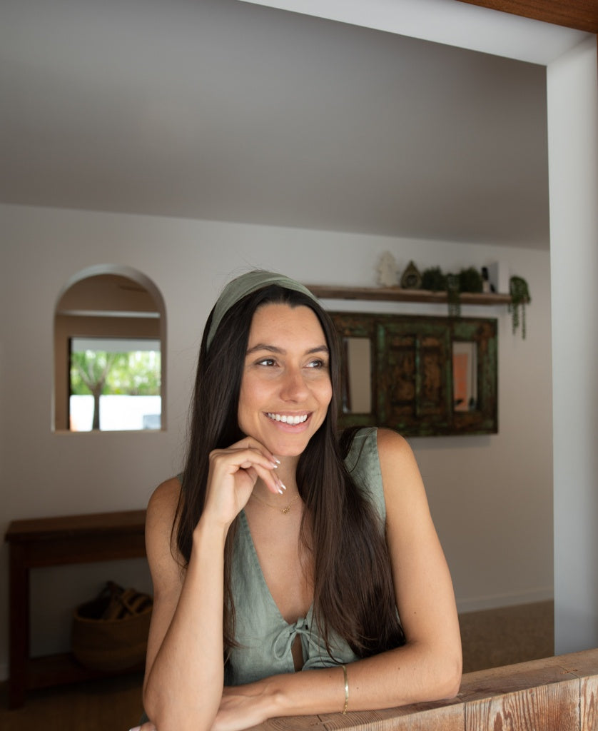 A woman with long dark hair smiles while leaning on a wooden surface in a bright room, wearing a Lazy Beach Head Scarf Linen in Dark Khaki.
