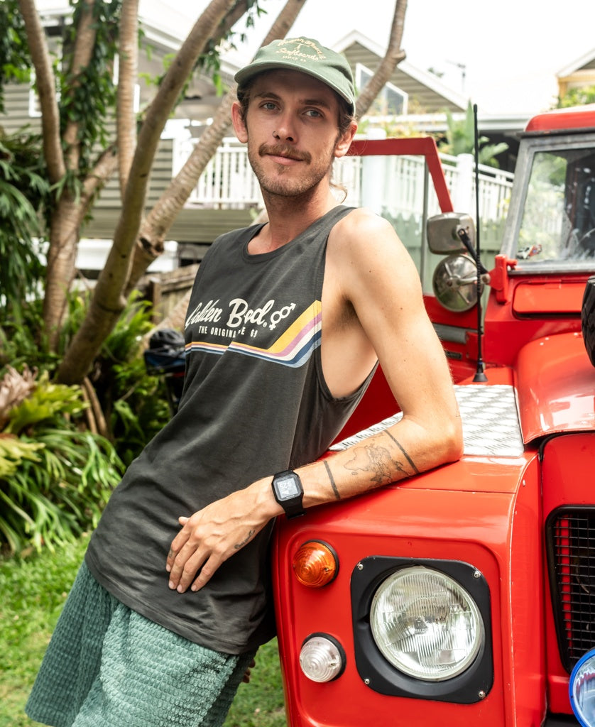 A man in a Golden Breed Retro Muscle Tank in Wash Black, paired with shorts and a cap, leans against a red off-road vehicle parked in a tree-lined yard with houses in the background.