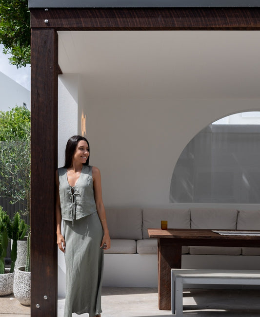 A woman wearing the Lazy Beach Pearl Maxi Skirt Linen in Dark Khaki stands by a wooden post in an outdoor seating area, pairing it with a sleeveless grey dress for a chic autumn look.