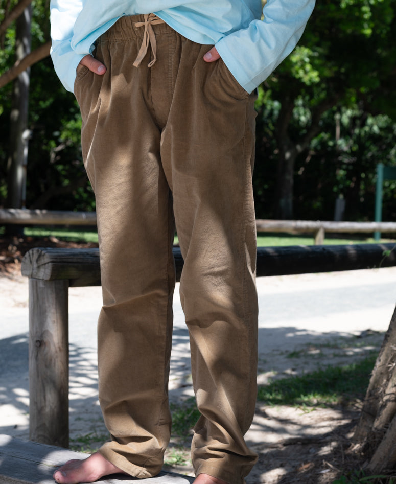 Person standing outdoors wearing Fred Stretch Cord Pant | Taupe with hands in pockets, paired with a light blue top, surrounded by trees and natural light.