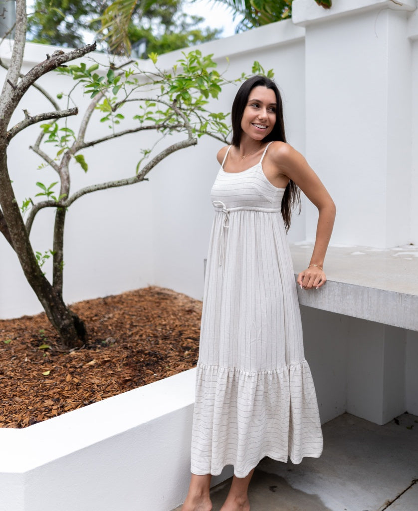 A woman stands barefoot outdoors near a white wall and small tree, wearing the Ali Maxi | Fine Stripe by Lazy Beach—a long, light-colored, sleeveless dress with elegant pinstripes perfect for easy summer style.
