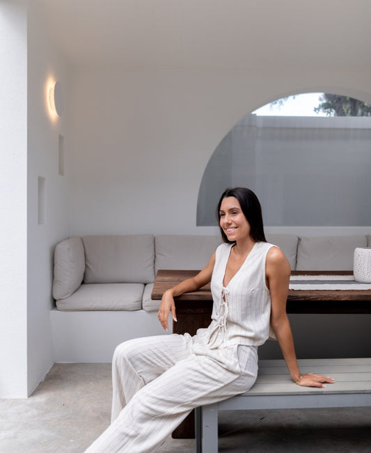 A woman in Lazy Beach’s Janine Pant | Fine Stripe and a light, sleeveless top sits on a bench in a modern, minimalistic room with built-in cushioned seating and a wooden table.