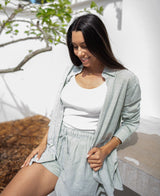 A woman wears the Judith Short in Olive Stripe by Lazy Beach, sitting on a concrete bench outdoors, smiling and looking down. A tree and white wall are behind her. Her outfit is handmade in Indonesia.