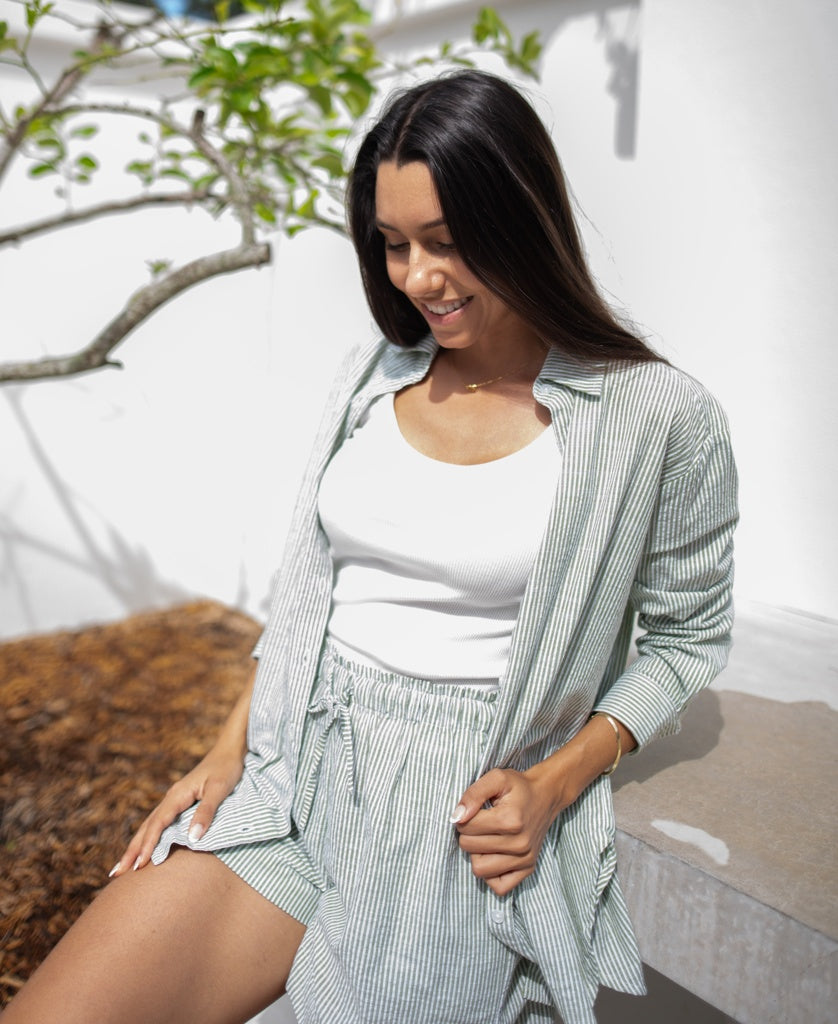 A woman wears the Judith Short in Olive Stripe by Lazy Beach, sitting on a concrete bench outdoors, smiling and looking down. A tree and white wall are behind her. Her outfit is handmade in Indonesia.