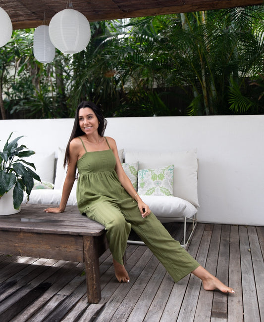 A woman in a Lazy Beach Celine | Khaki Stripe jumpsuit sits barefoot on a wooden bench in an outdoor lounge filled with plants, white cushions, and hanging lanterns.