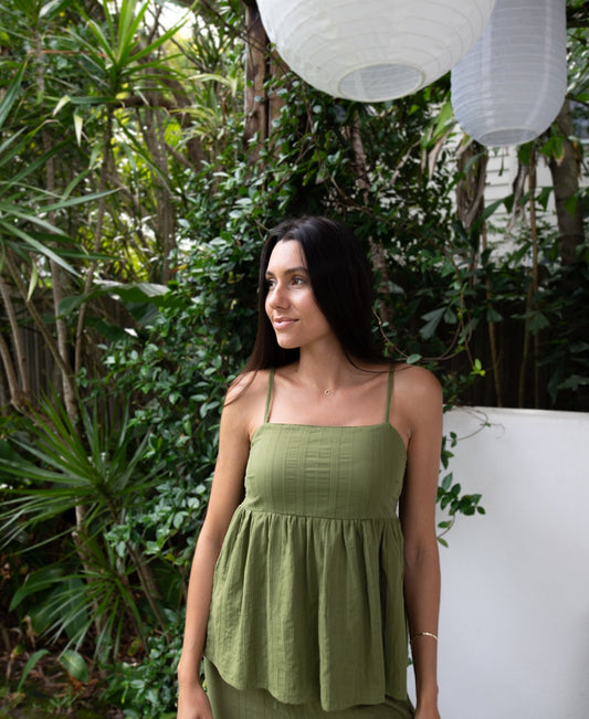 A woman in a Lazy Beach Celine Khaki Stripe sleeveless top stands outdoors near green plants and white paper lanterns, looking to the side.