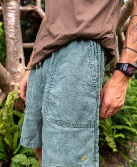 A person wears Golden Breeds Linen Short in Khaki, standing outdoors from mid-torso to knee, with lush greenery and tree branches in the background.