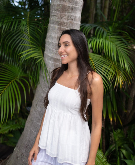 A woman with long brown hair wears the Lazy Beach Celine | White Stripe top and smiles outdoors in front of a palm tree and lush green foliage.
