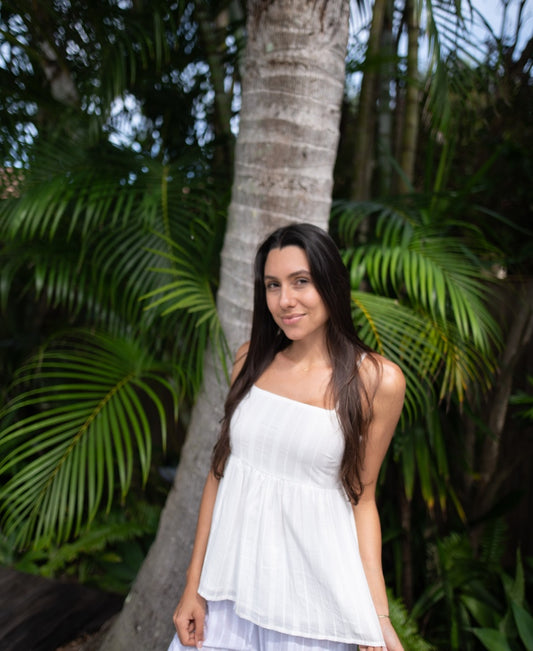 A woman wears the Lazy Beach Celine | White Stripe top, handmade in Bali from 100% cotton, standing outdoors by a palm tree and lush green foliage.