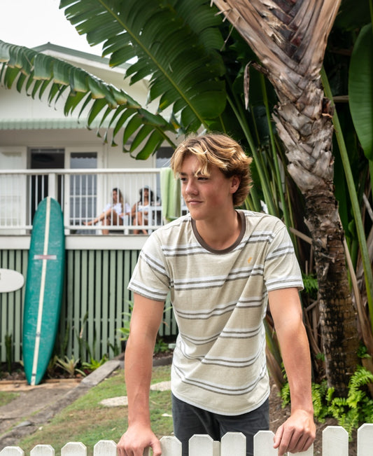 A young man in the Golden Breed Bay Stripe Tee in Mocha stands by a white picket fence, with two surfboards and people on a house porch in the background, surrounded by tropical plants.