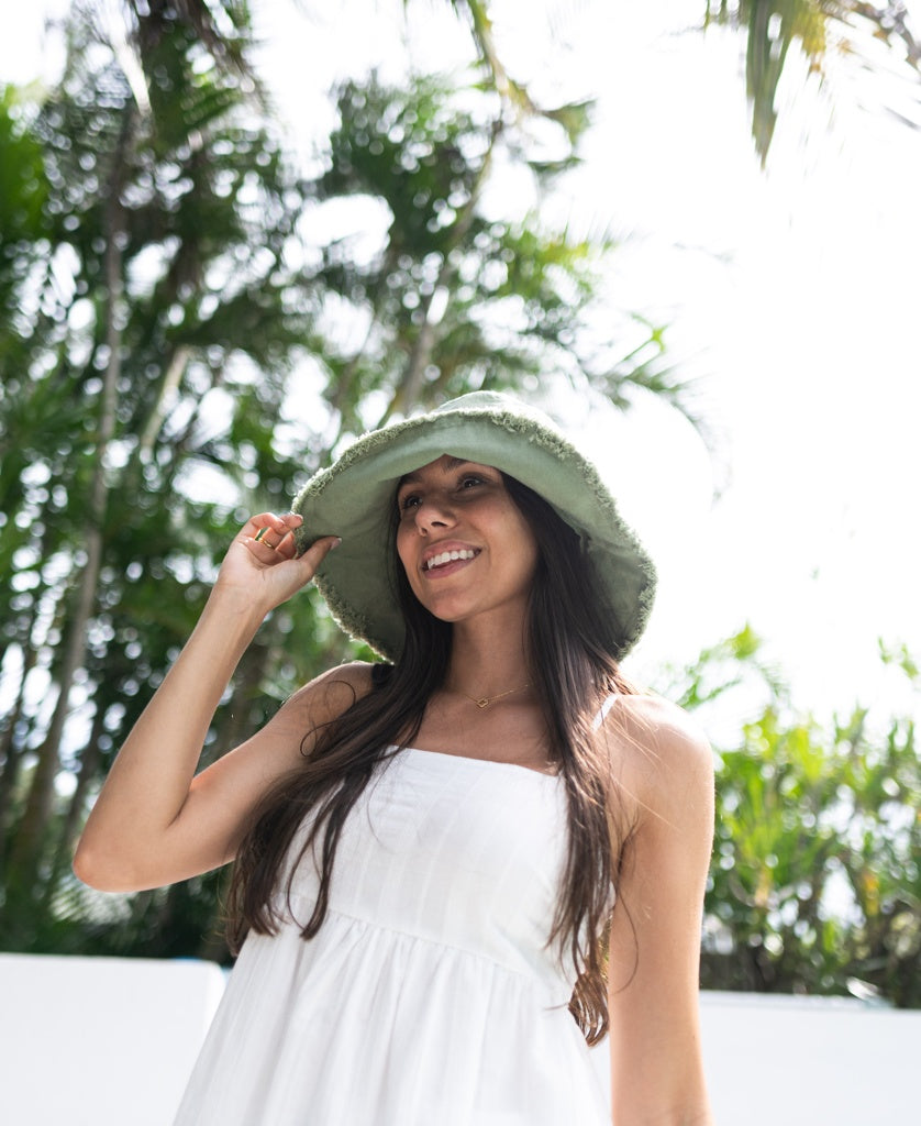 A woman wearing the Lazy Beach Halo Bucket Hat in Olive stands outdoors, smiling with palm trees and sunlight in the background.
