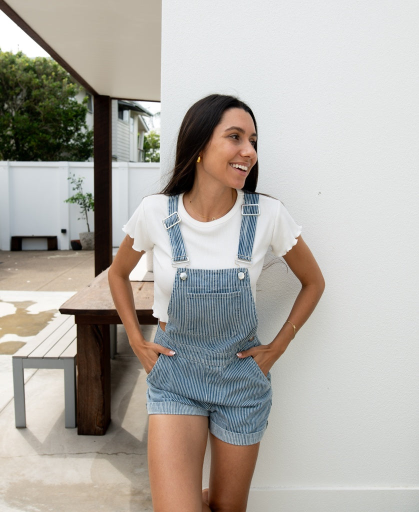 A woman in Golden Breed’s Opal Short Overall in Light Blue Stripe, made from 100% cotton with adjustable straps, wears a white t-shirt as she stands outside, smiling and leaning against a white wall.