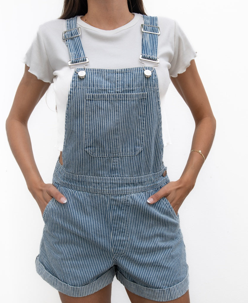 A person wearing Golden Breed’s Opal Short Overall in Light Blue Stripe—100% cotton with adjustable straps—and a white short-sleeve shirt, hands in pockets, stands against a plain white background.