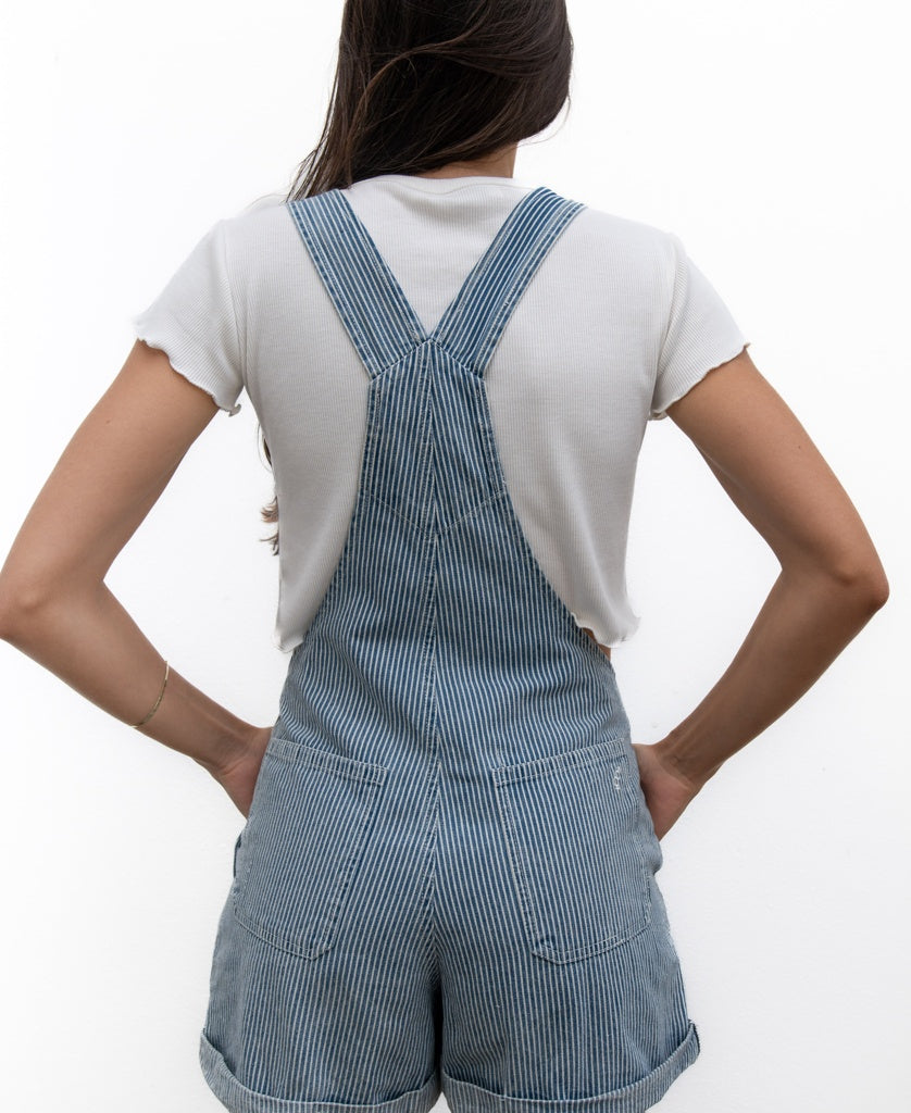 A person stands with hands on hips against a white background, wearing Golden Breeds Opal Short Overall in light blue stripe—100% cotton with adjustable straps—paired with a white short-sleeve shirt.