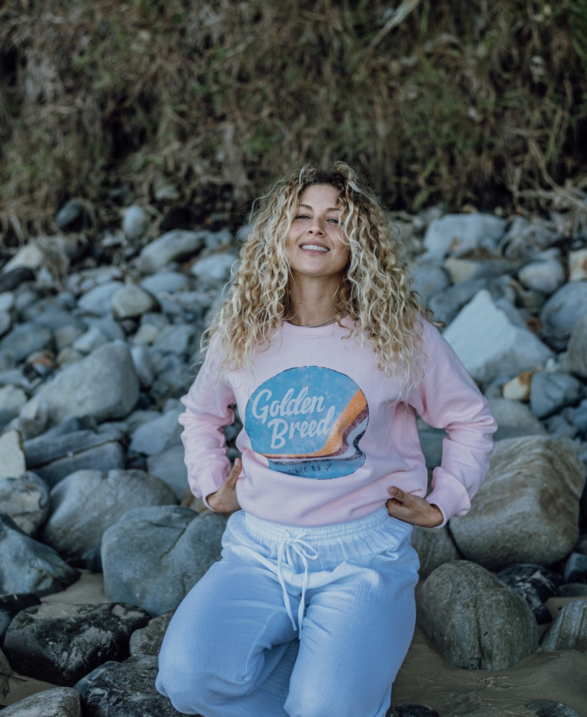 A woman with curly blonde hair, dressed in the Golden Breed Vintage Sweat | Pale Pink and white pants, kneels on rocky ground near a hillside.