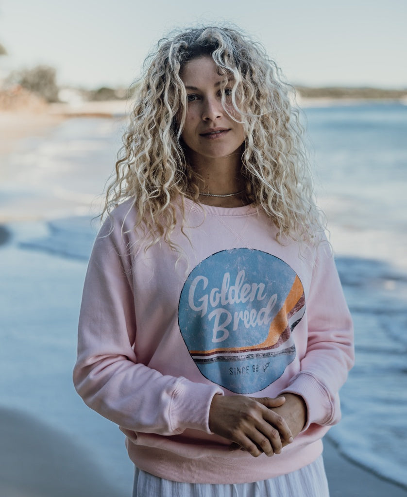A woman with curly blonde hair stands on a beach wearing the Golden Breed Vintage Sweat in pale pink, featuring a retro circular logo graphic.