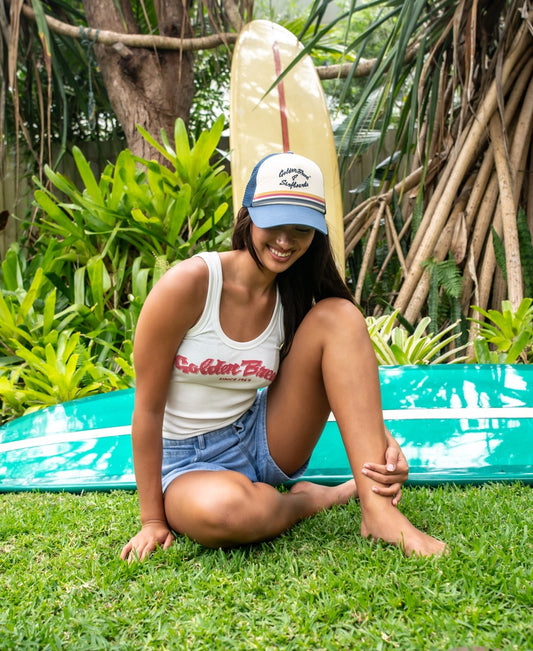 A woman sits on grass before two surfboards, wearing a tank top, denim shorts, and the Golden Breed GoJo Trucker Cap in Dark Denim with an adjustable strap, surrounded by lush greenery and tree roots.