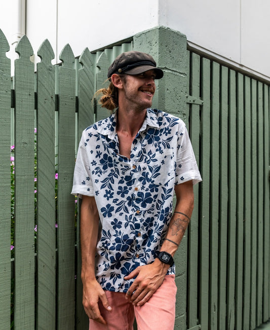 A man wearing an oversized relaxed fit Golden Breed Vomo Floral Shirt, pink shorts, and a black cap stands in front of a green fence, smiling and looking to the side.