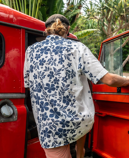 Wearing a Golden Breed Vomo Floral Shirt with blue floral patterns and an oversized relaxed fit, along with a cap, a person steps into a red vehicle amid greenery.