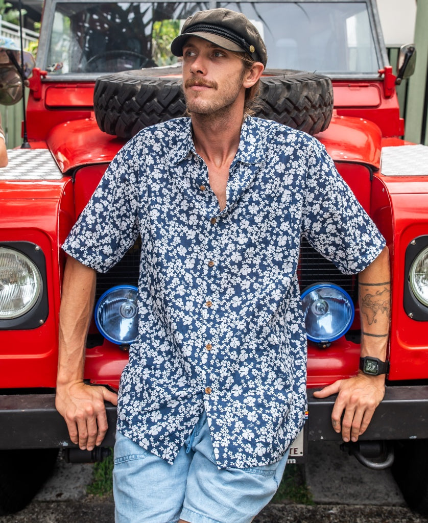 A man in a Golden Breed Liberty Floral Shirt | Vintage Navy and cap leans against the front of a red vehicle with a spare tire on its hood.