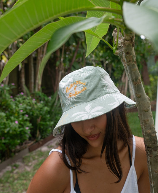 A woman stands outdoors near a tree, wearing the Golden Breed Chelsea Bucket Hat in Sage. The lightweight cotton linen fabric keeps her comfortable as she enjoys the fresh air surrounded by greenery.