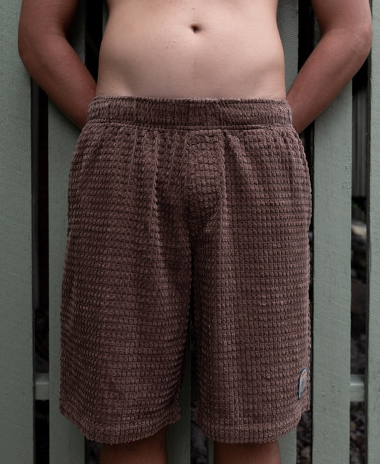 A person wears Golden Breed El Solito Walkshort in Wash Brown, featuring an elastic waist and chunky cotton cord, shown from waist to mid-thigh before a light green wooden fence.