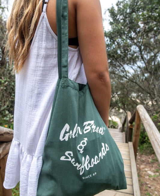 Wearing a white sleeveless dress, a person carries the Golden Breed Surf Logo Tote Bag in Ivy Green while standing on wooden steps outdoors near trees.