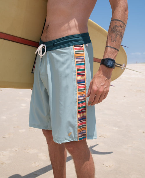 A person wearing Golden Breed’s Sunny Island Boardie in Sage, featuring a colorful striped side panel, stands on the beach with a surfboard, capturing an authentic California surf vibe.