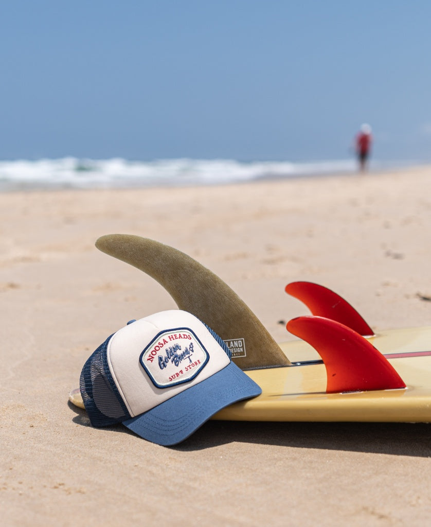 A Golden Breed Noosa Trucker Cap | Dark Denim sits on the sand beside a surfboard with red fins at Byron Bay’s shoreline, with waves and a person visible in the background.