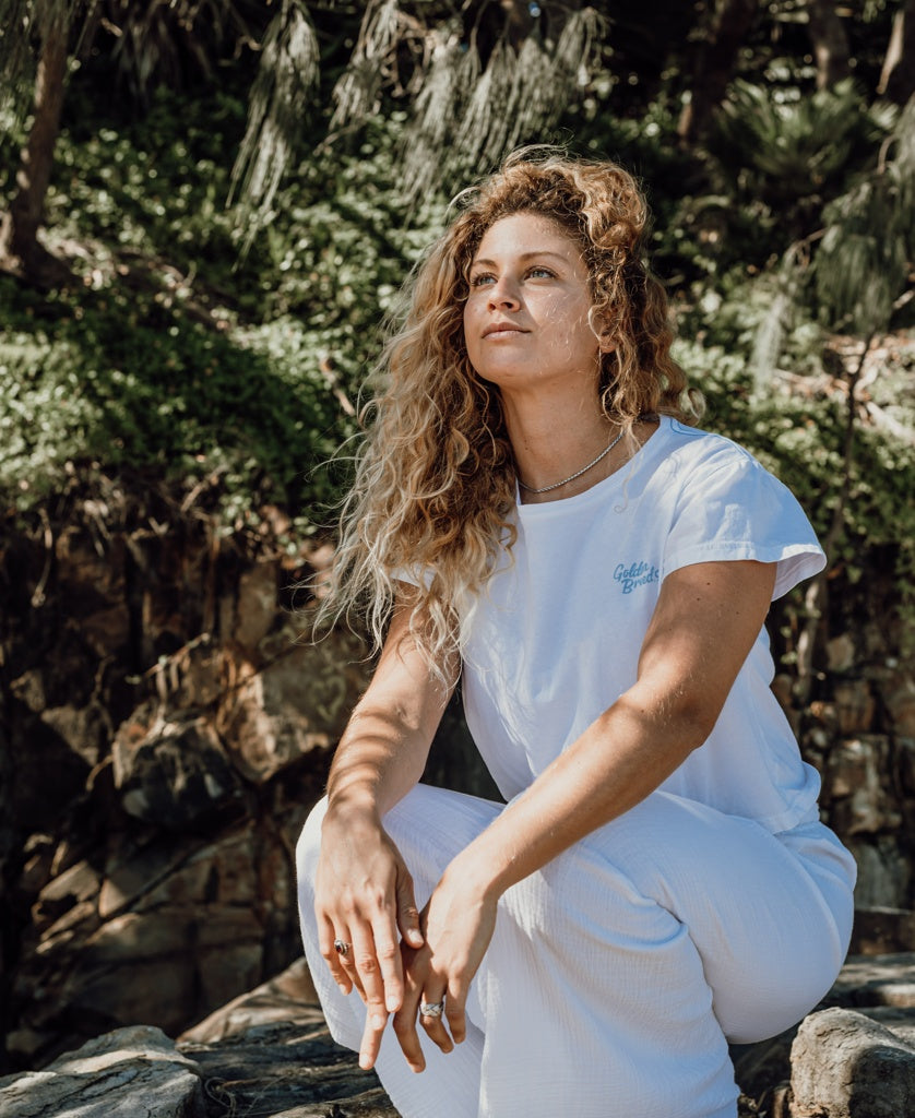 A woman with curly hair wears the Golden Breed Hattie Crop Tee in white and pants, sitting outdoors on rocks and gazing thoughtfully into the distance, with greenery in the background.