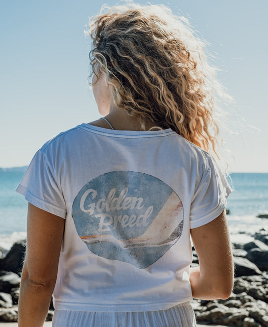 A person with curly blonde hair stands on a rocky beach, facing the sea, wearing the Golden Breed Hattie Crop Tee in white with branding on the back.