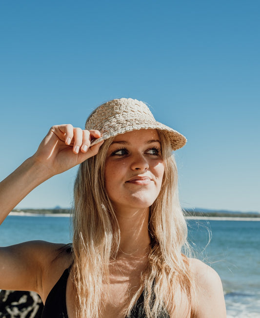 A woman with long blonde hair wears the Lazy Beach Palm Visor | Natural, paired with a black top, standing near the water under a blue sky.