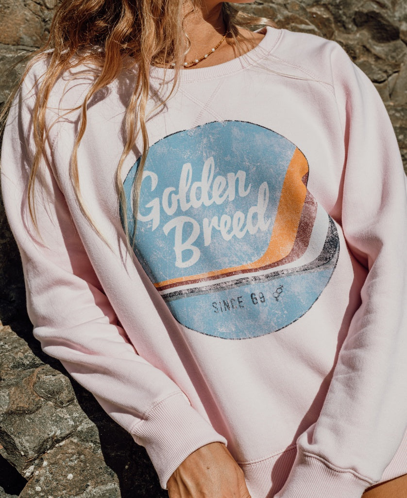 A person stands outdoors against rocky terrain, wearing the Golden Breed Vintage Sweat in pale pink featuring a retro logo, made from 100% cotton fleece.