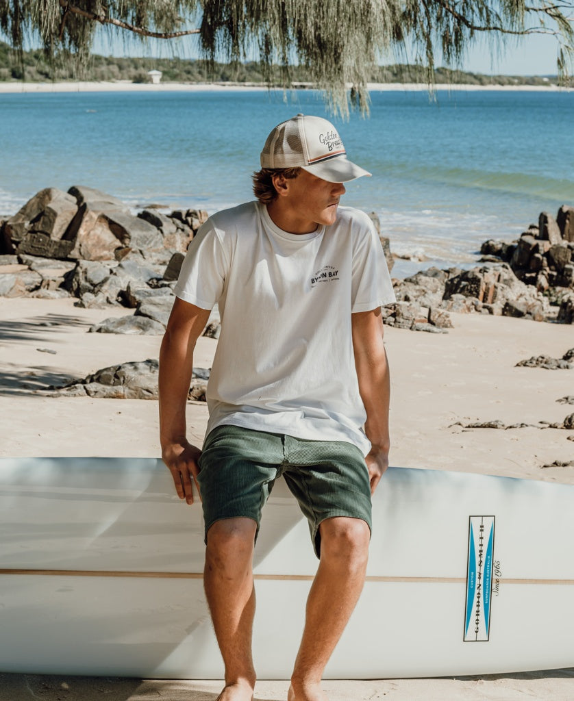 A man wearing a white t-shirt, green shorts, and the Golden Breed Retro Trucker Cap in Off White sits on a surfboard on a sandy beach near rocks, with the ocean and trees in the background.