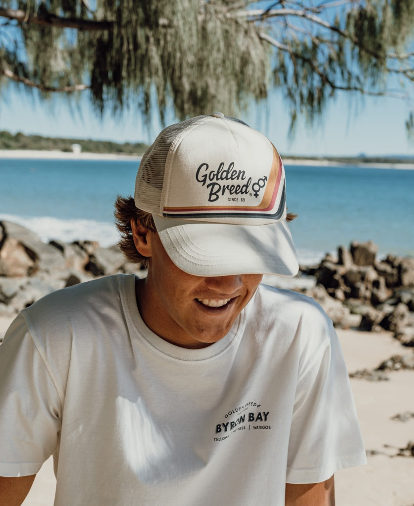 Wearing a Golden Breed Retro Trucker Cap in Off White with an adjustable strap and a Byron Bay t-shirt, a person stands on a beach surrounded by rocks, trees, and water.
