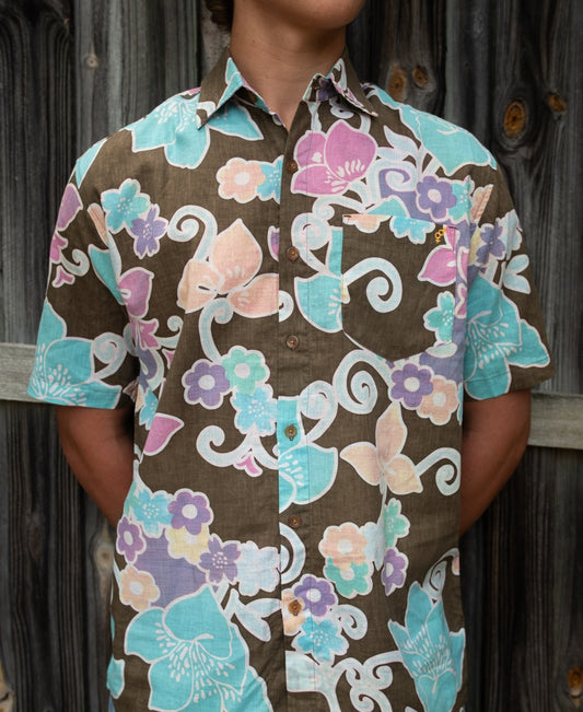 A person wearing a short-sleeved Golden Breed Batik Shirt in Wash Brown stands in front of a wooden fence with their arms behind their back.