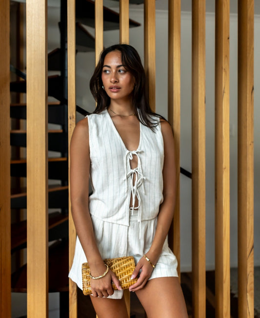 A woman wearing the Lazy Beach Rennie Vest in Off White Fine Pinstripe, hand made in Bali, holds a woven clutch while standing indoors before vertical wooden slats and a staircase.