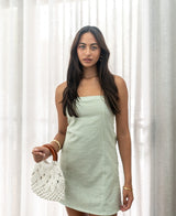A woman wearing the Lazy Beach Victoria Dress in Olive Stripe stands before sheer curtains, holding a white woven handbag with brown handles.