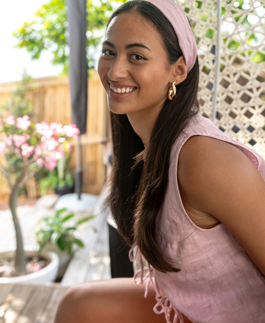 A woman with long dark hair smiles while sitting outdoors, wearing the Lazy Beach Head Scarf in blush linen and a sleeveless top, with plants in the background.