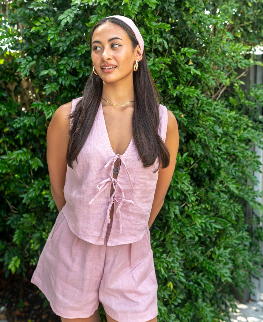 A woman stands before green bushes, hands behind her back, wearing the Lazy Beach Rennie Vest in Blush Linen—handmade in Bali for effortless warm-weather style.
