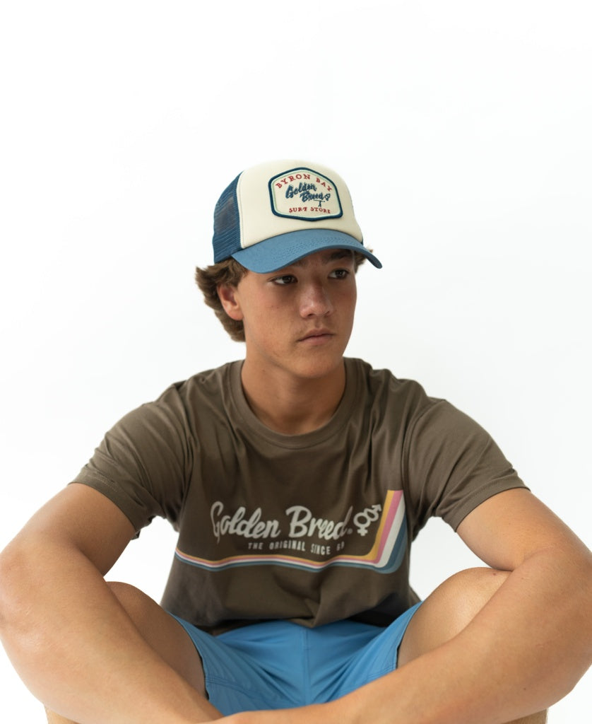 Teen boy in a Golden Breed t-shirt, dark denim blue shorts, and a Golden Breed Byron Trucker Cap in Dark Denim, sits against a plain white background.