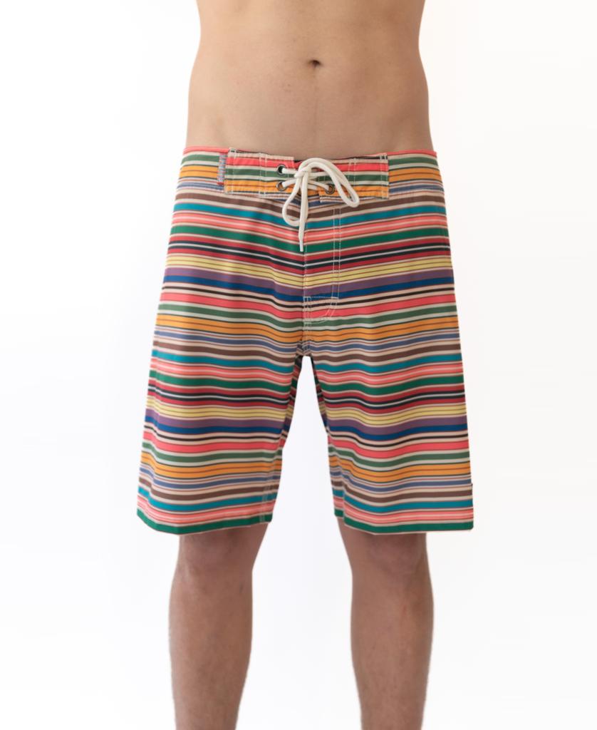 A person wears Golden Breeds Daze Boardie | Stripe, multicolored striped boardshorts with a white drawstring made from recycled polyester, standing against a plain white background with torso partially out of frame.